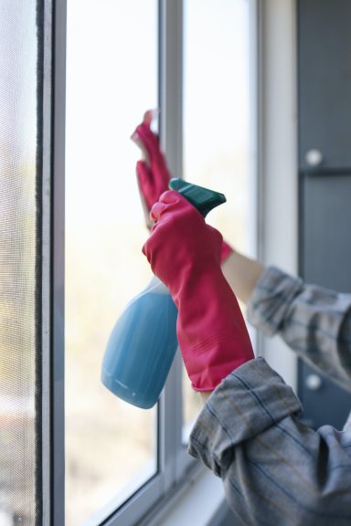 Manos con guantes rosas sosteniendo un spray limpiador junto a una ventana.