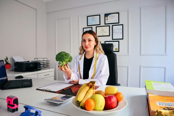 Mujer con bata de médico sosteniendo brócoli en un consultorio con frutas en la mesa.